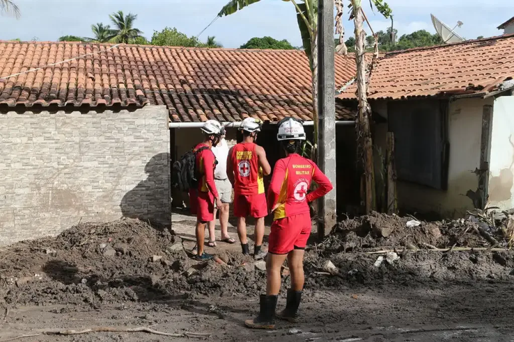 Três bombeiros na frente de uma casa atingida pela chuva, com muita terra na frente da casa.