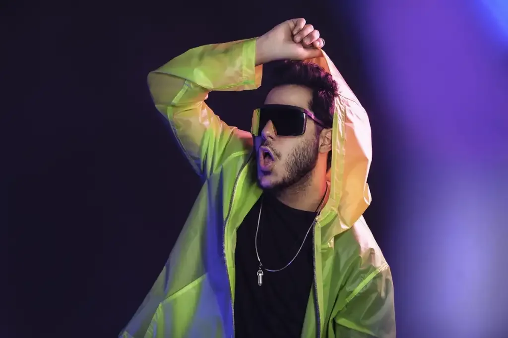 Homem de camisa preta, capa de chuva amarerela e óculos escuros e fundo preto e roxo.