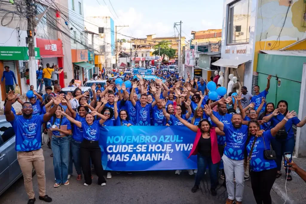 Rua lotada de homens vestidos de camisa azul fazendo caminhada com uma faixa enorme sobre a campanha novembro azul