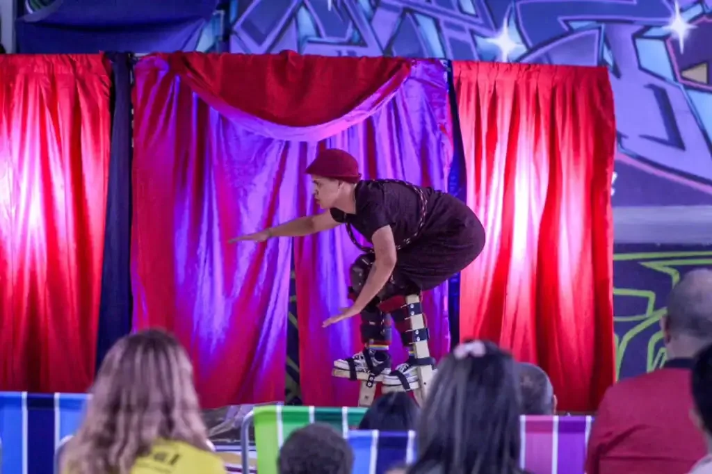 homem andando encurvado na perna de pau em um palco com corninas roxa e vermelha, algumas pessoas assistindo