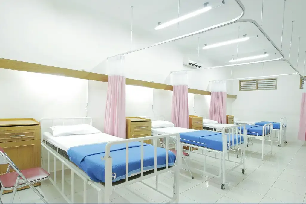 Sala de hospital com trÊs leitos de lençois branco e azul e cortimas rosa fechadas.